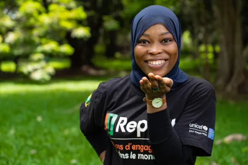 A young girl wearing a hijab and U-report T-shirt smiles at the camera while standing outdoors.