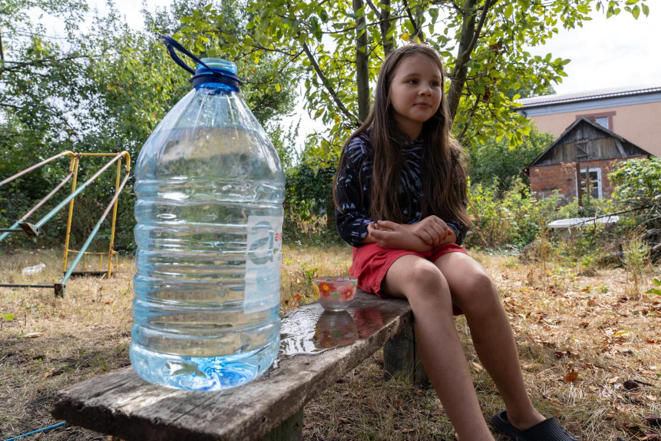 A girl sits outside on a bench with a large bottle of water beside her.