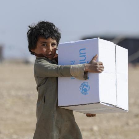A child holds a UNICEF box, in the background out of focus you can see tents