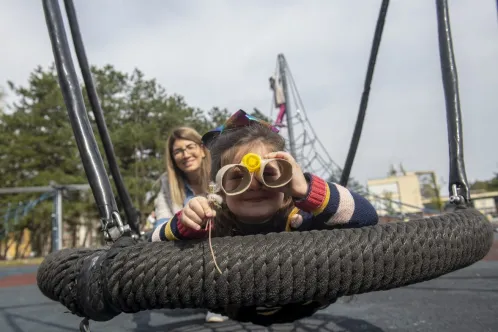 A young girl sitting on a playground swing looks through homemade binoculars. Her mother watches on in the background.