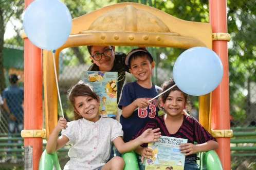 Four children stand together on a play structure and smile while holding balloons and reading materials. 