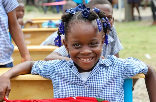 In Haiti, a girl smiles at the camera while sitting with her arms resting on a school desk, in an outdoor classroom.