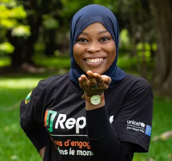 A young girl wearing a hijab and U-report T-shirt smiles at the camera while standing outdoors.