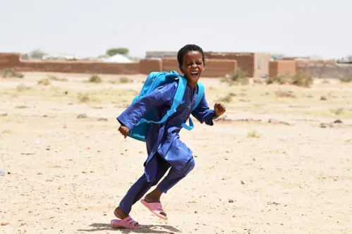 A young boy wearing a blue UNICEF backpack runs while smiling at the camera.