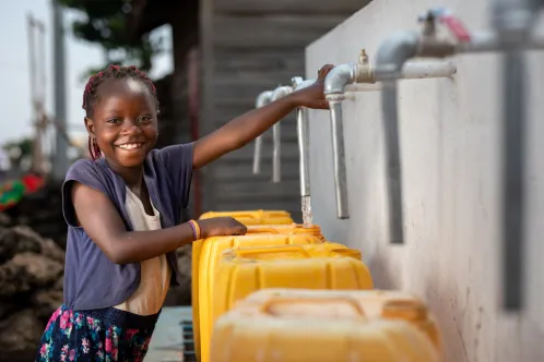 A girl smiles at the camera while filling a yellow water jug at an outdoor tap.