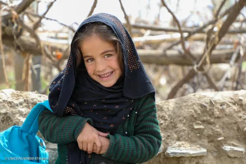  In Afghanistan, a young girl smiles at the camera, with the blue strap of a UNICEF backpack over her arm.