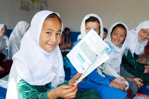 In a classroom, a girl wearing a white headscarf holds an open book and smiles at the camera.