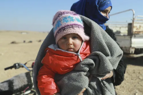 Une femme tient dans ses bras un jeune enfant portant des vêtements d’hiver et enveloppé dans une couverture grise.