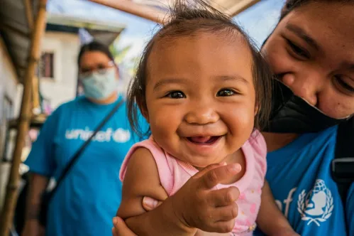 Gros plan sur une fillette qui sourit tandis qu’un membre du personnel de l’UNICEF se tient en arrière-plan. 