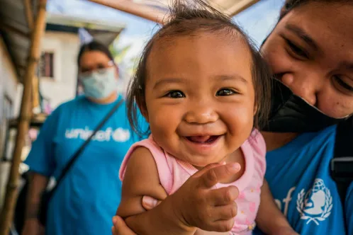 A young child being held by a UNICEF worker smiles at the camera.