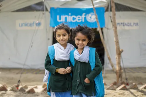 Two young girls wearing blue UNICEF backpacks smile at the camera while standing in front of a UNICEF-funded Temporary Learning Centre