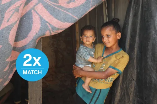 A girl stands at the opening of a makeshift tent shelter holding a toddler on her right hip.