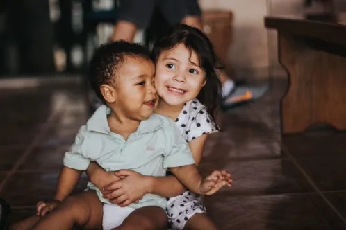 A toddler sits on the floor in a diaper, smiling, as a female child gives him a hug from behind. 