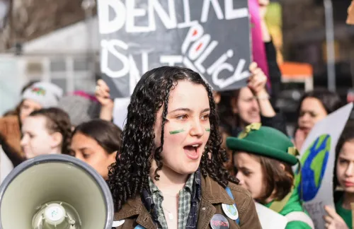 Olivia, 16, wears green face paint and carries a megaphone at a climate protest, surrounded by other protestors with signs.