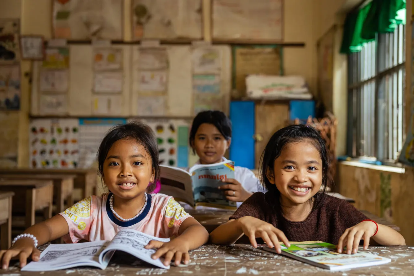 Dans une salle de classe au Cambodge, trois filles assises à leur pupitre avec leurs cahiers ouverts sourient à l’objectif.