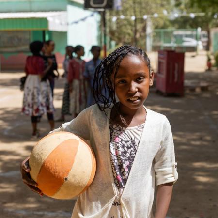 A child stands holding a basketball while smiling at the camera in an outdoor setting with other children standing in the background