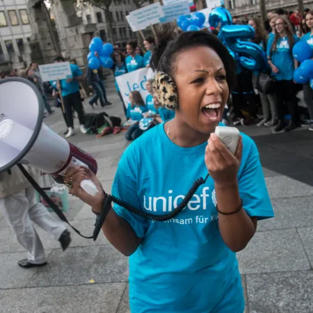 A girl wearing a blue UNCIEF tshirt speaks into a speakerphone, behind her others can be seen standing in a line also wearing UNICEF tshirts.