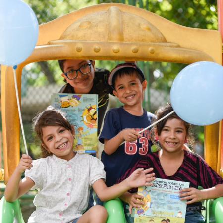 Four children stand together on a play structure and smile while holding balloons and reading materials.