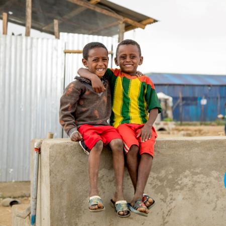 Two children sit on a concrete wall with their arms around each other’s shoulders and smiling at the camera.