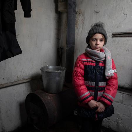 In Ukraine, a girl wearing winter clothes stands beside a wood-burning stove.