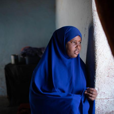 A girl wearing a blue headscarf look out of a window. A curtain is silhouetted in the foreground on the right side of the photo. 