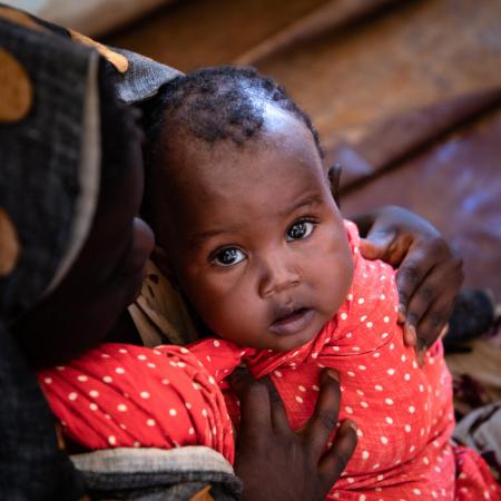 A child wrapped in a red polka dotted blanket looks up to the camera. he is held by his mother whose face is cast down.