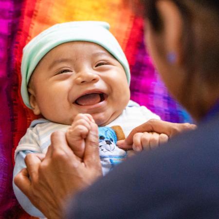 A woman plays with a baby lying on its back, she is holding his hands and the baby is smiling.