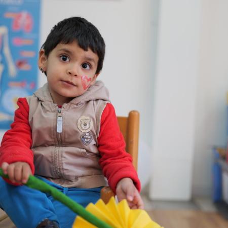 A child with paint on his face, in a classroom-like setting sits on a chair and looks directly into the camera.