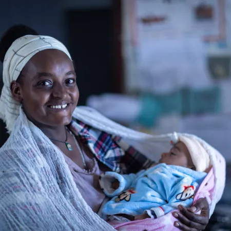 A woman looks at the camera smiling while holding a sleeping baby.