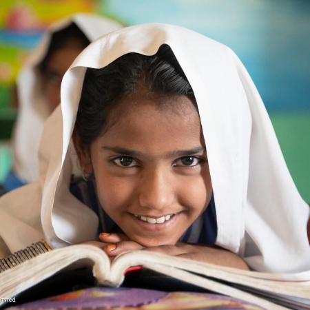 Dans une salle de classe au Pakistan, une jeune fille assise à son pupitre sourit, un livre ouvert posé devant elle. 