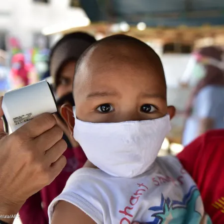A child wearing a mask looks at the camera as a woman behind him checks his temperature with a digital thermometer. This photo was taken in Thailand in 2020.