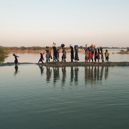 A group of people, including women and children, walk with water containers on their heads along a narrow, muddy path flanked by water in a rural setting.