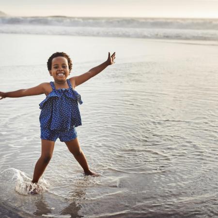 A young girl in stands in a body of water with her arms held wide smiling at the camera as the sun sets to the side of the photo