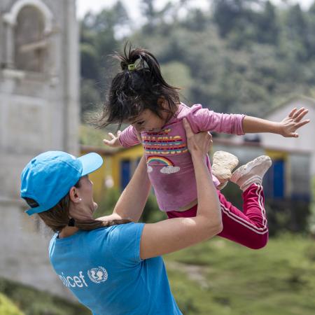 A woman, a UNICEF Staff member, raises a child high above her head, capturing a delightful moment of connection and joy in their interaction.