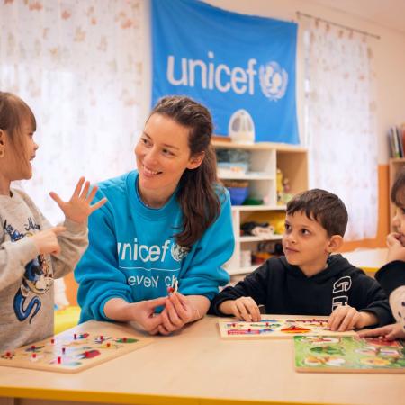 Two girls and a boy play around a table with a woman wearing a blue UNICEF sweater. 