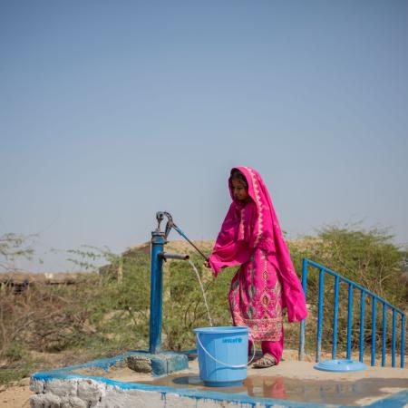 In a bright outdoor setting an adolescent stands and smiles while working the lever on a water pump as water pours from a spout into a bucket.