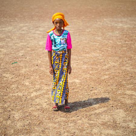 Une jeune fille se tient debout sur une terre aride, portant un chandail rose, une jupe à motifs, un foulard orange et des sandales.