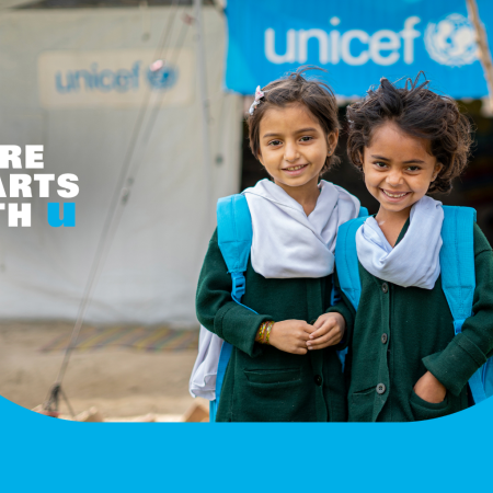 Two young girls wearing blue UNICEF backpacks smile at the camera while standing in front of a UNICEF-funded Temporary Learning Centre.