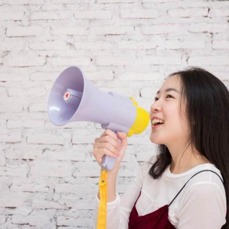 A young girl, standing alone in front of a white wall, smiles while holding up a megaphone.