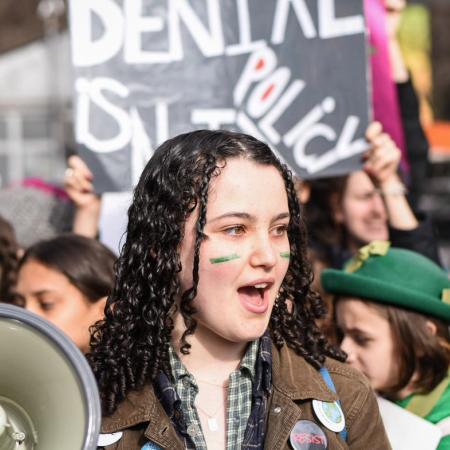 Olivia, 16, wears green face paint and carries a megaphone at a climate protest, surrounded by other protestors with signs.